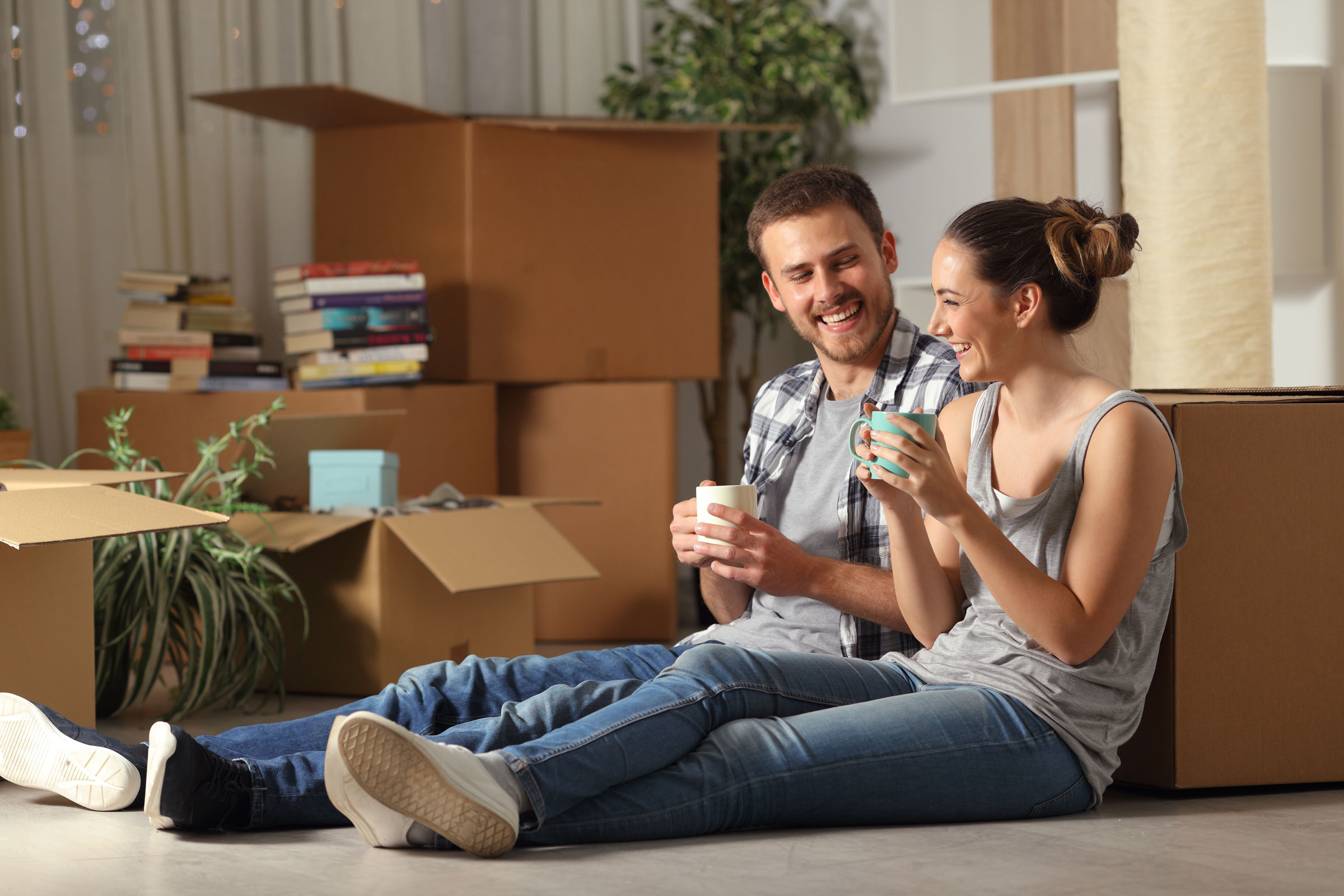 A couple sitting on the floor among a pile of open boxes, drinking from mugs and laughing.