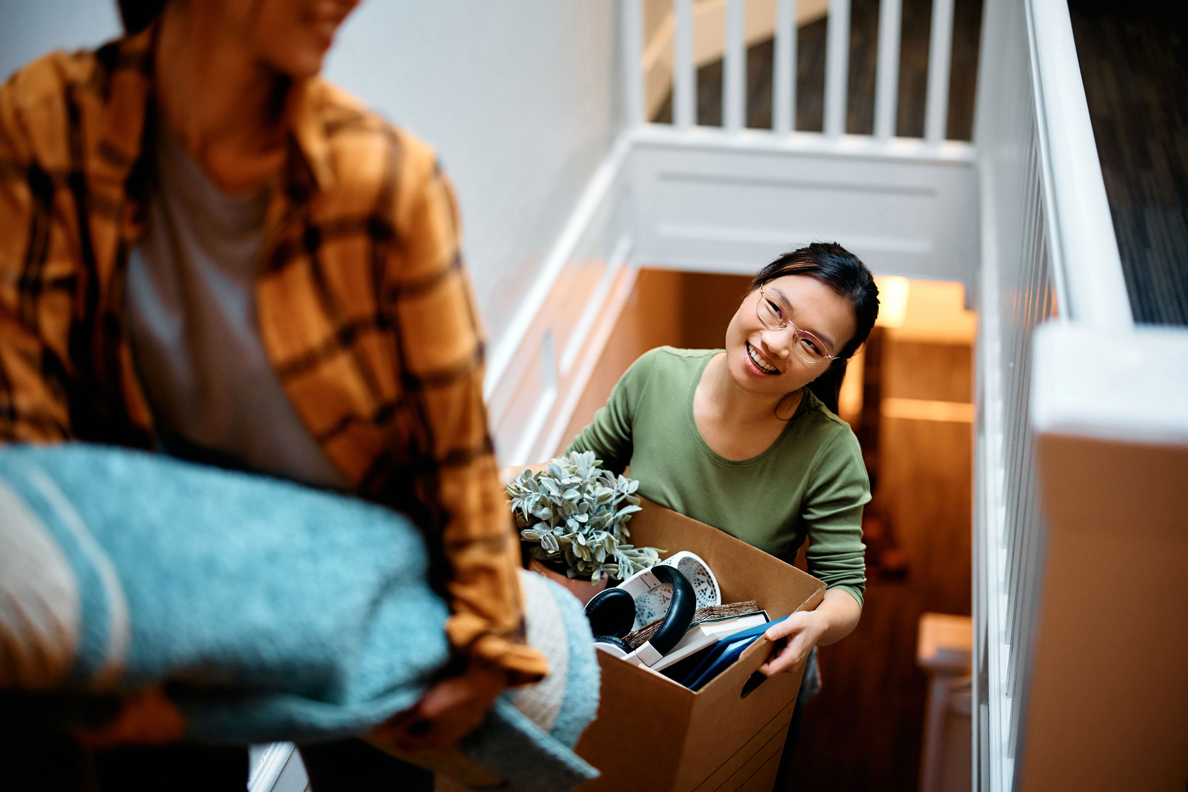 Woman walking up the stairs holding a box full of random items, smiling at the person in front of her.